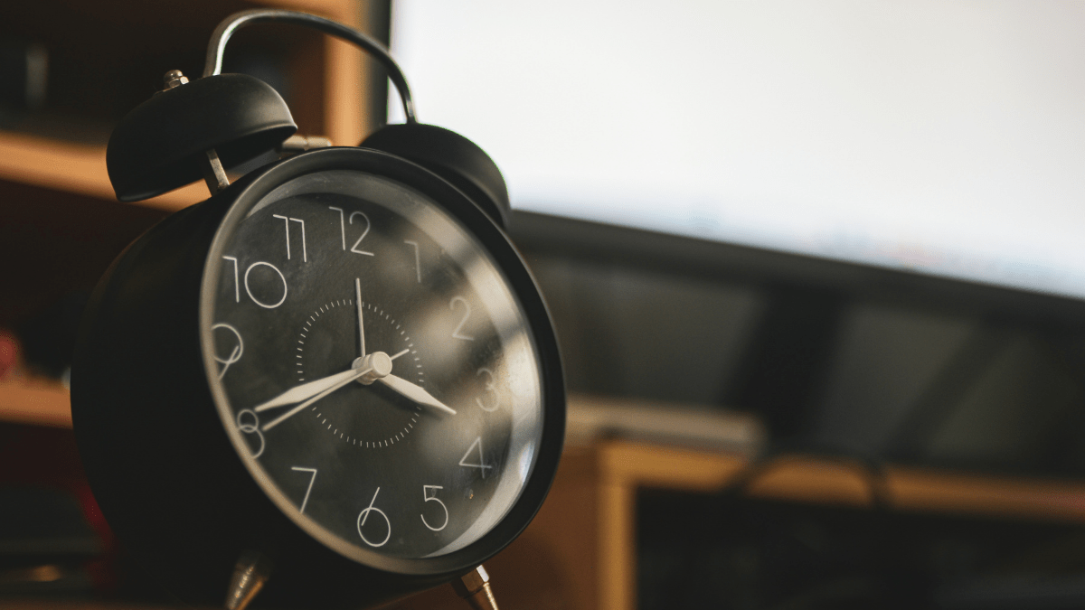 Black analog alarm clock showing 4:00 on a bedside table in soft morning light.