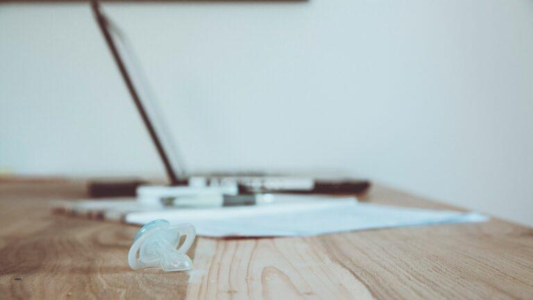A baby pacifier resting on a desk in front of a laptop and paperwork, symbolising the mental load of balancing caregiving and work.