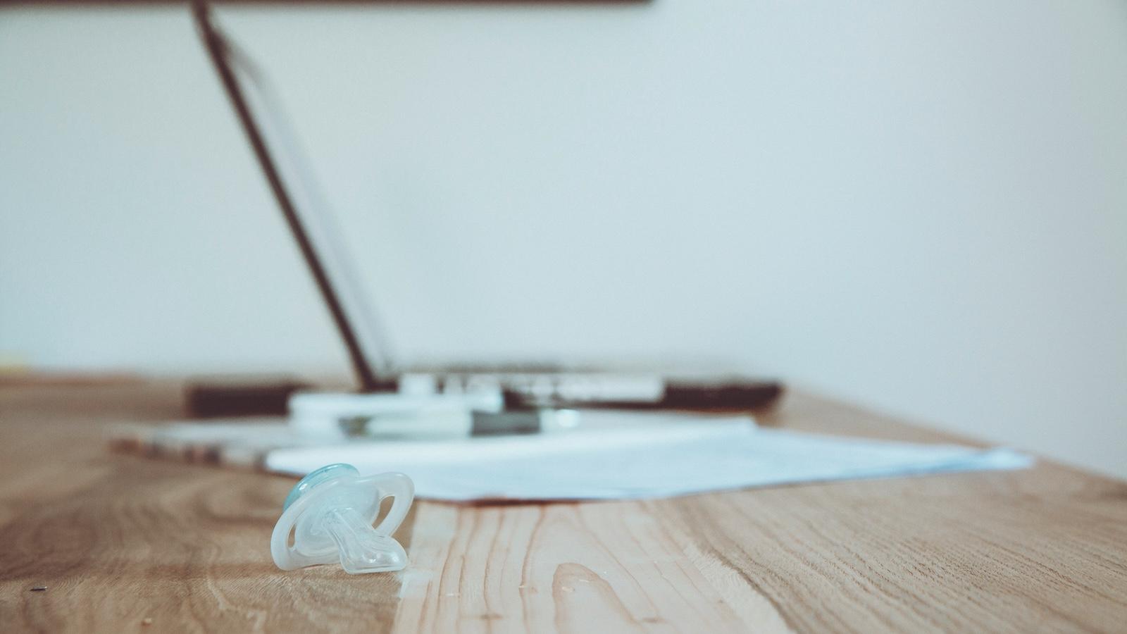 A baby pacifier resting on a desk in front of a laptop and paperwork, symbolising the mental load of balancing caregiving and work.