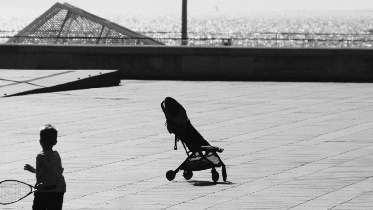 Black-and-white photo of a young child running across a large open plaza while holding a tennis racket, with an empty stroller nearby and the sea glinting in the background.
