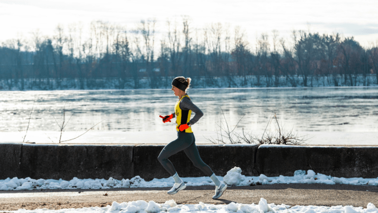 A person wearing winter running gear jogs along a snowy riverside path, with a frozen or partially frozen river and leafless trees in the background.