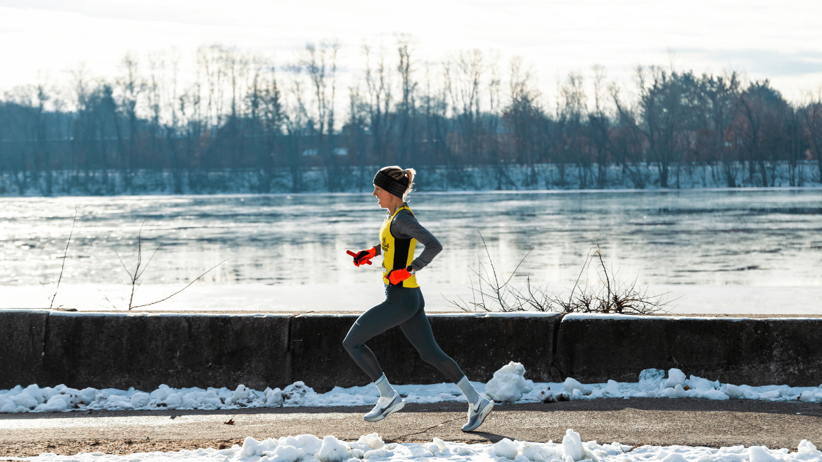 A person wearing winter running gear jogs along a snowy riverside path, with a frozen or partially frozen river and leafless trees in the background.
