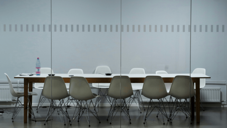 Empty meeting room seen through glass partition, grey walls, long wooden table surrounded by empty white chairs.