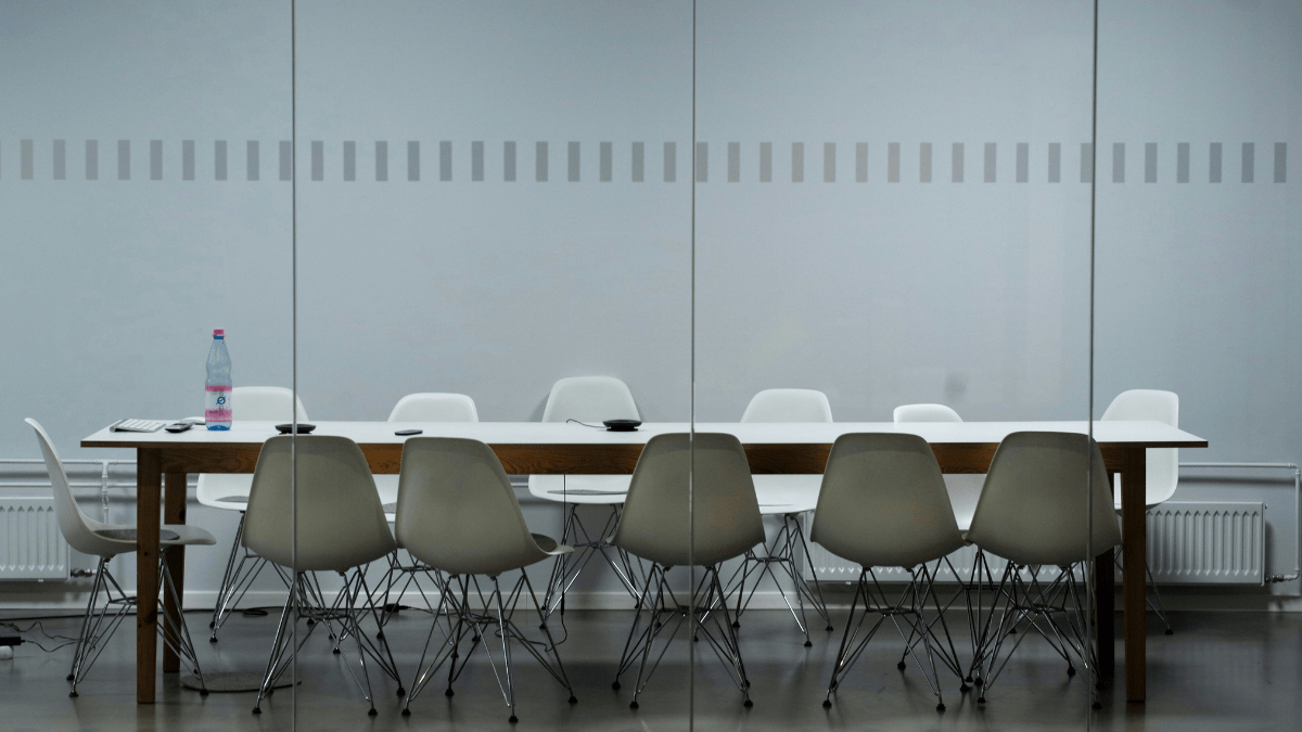 Empty meeting room seen through glass partition, grey walls, long wooden table surrounded by empty white chairs.