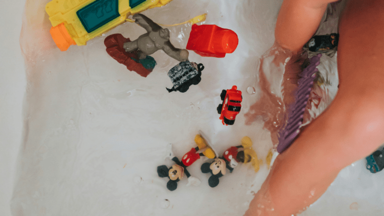 Close-up of a child’s legs in a bath with water and bubbles, surrounded by small toy figures and vehicles floating in the tub.