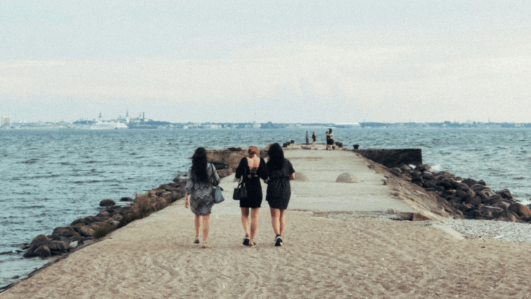 Three women walking together along a stone jetty toward a distant city across the water